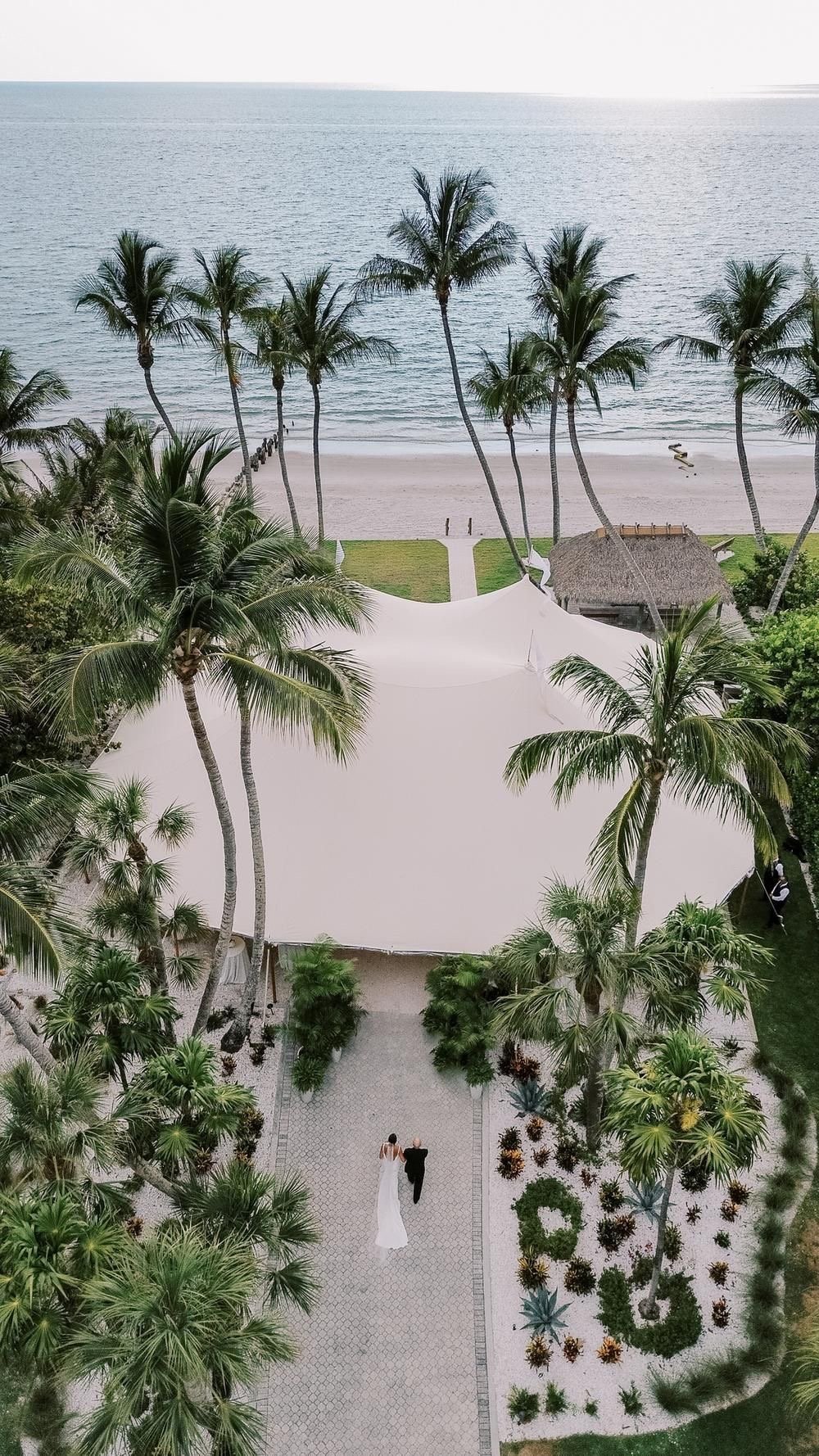 Aerial view of beachfront property with palm trees, manicured gardens, sandy beach, and couple walking near thatched-roof structure by ocean