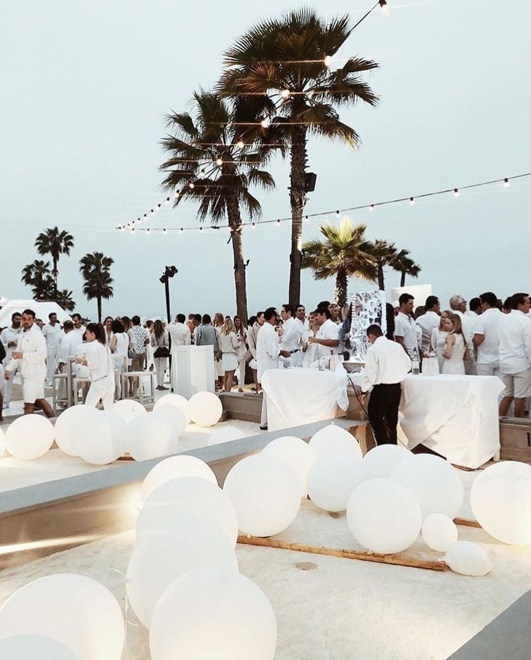 Outdoor beach event with guests in white attire, palm trees, string lights, and white balloon decorations on sand