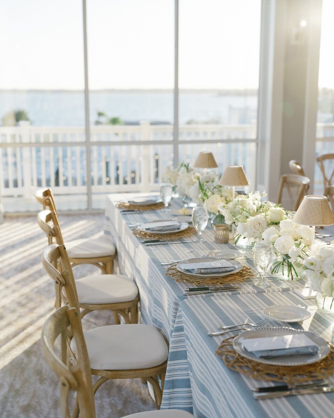 Elegant coastal dining table with white place settings, blue striped runner, white floral centerpieces, and ocean view through floor-to-ceiling windows