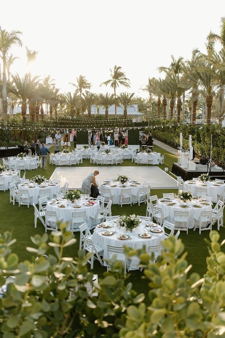 Elegant outdoor wedding reception with white round tables on a manicured lawn, palm trees, and ocean view in background
