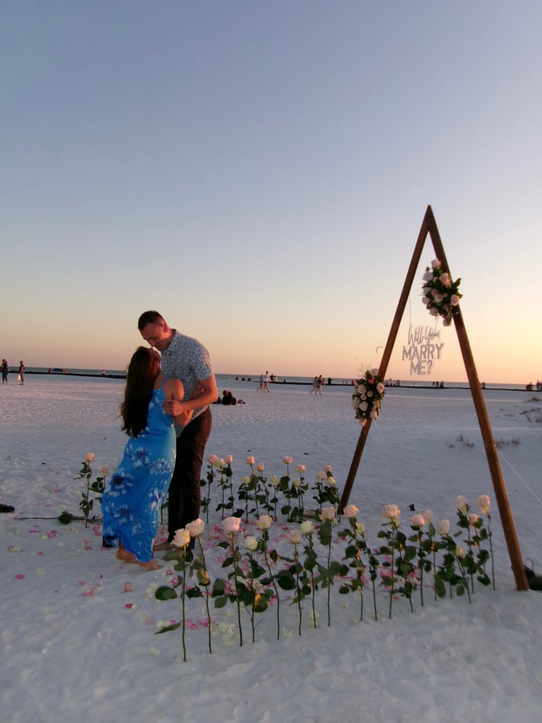 Couple embracing on beach at sunset with wooden arch decorated with flowers and marry sign, surrounded by rose bouquets in sand